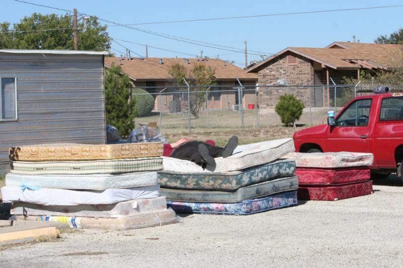 Rows of discarded mattresses stacked on pavement near a red pickup truck and residential buildings