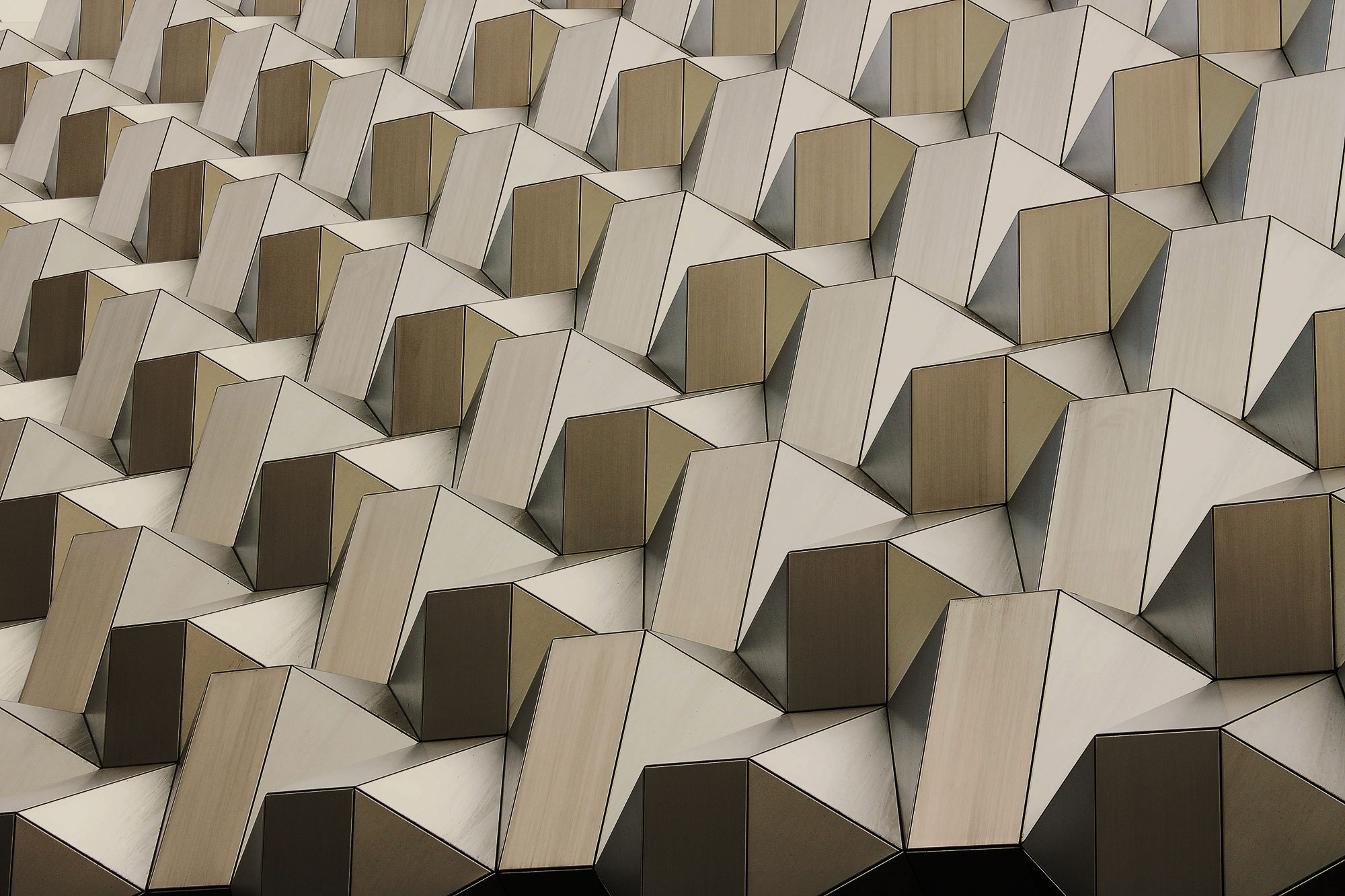 Repeating geometric pattern of angular metallic panels in gold, brown, and cream tones on a building facade